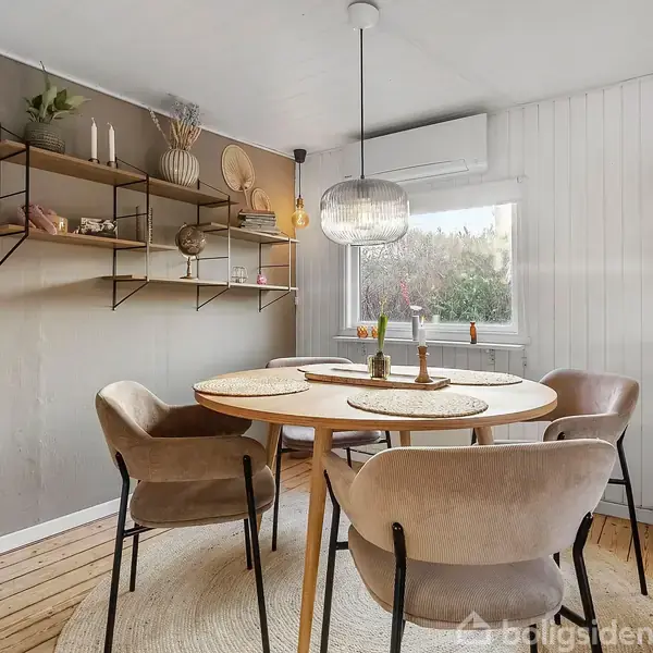 Rundt spisebord med fire polstrede stole står i en lys stue. Hylder med dekorationer hænger på væggen. Hængelampe over bordet og stort vindue til højre med udsigt.