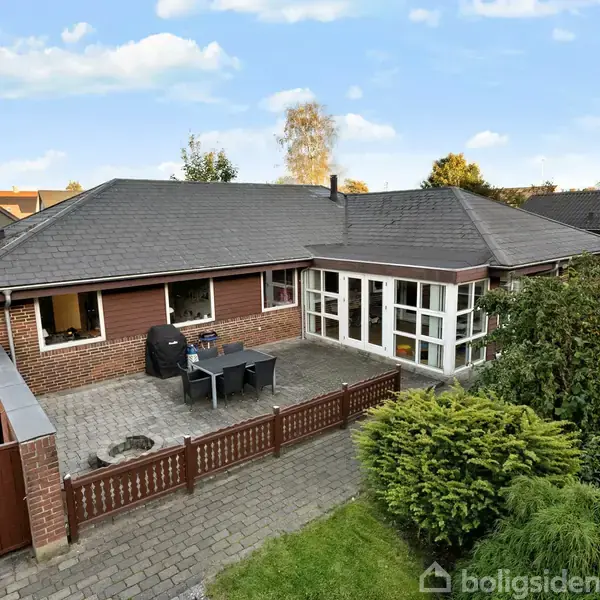 En murstensvilla står stille i en rolig have. En terrasse med bord og grill ses ved siden af huset, omkranset af grønne buske under en lys, skyet himmel.