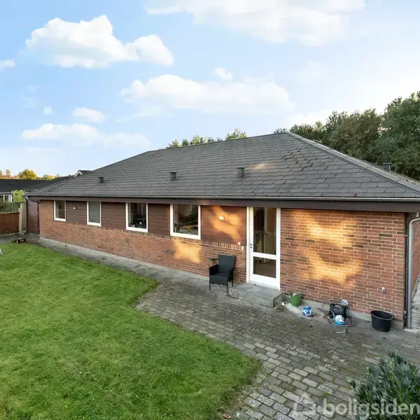 Et enfamiliehus af mursten står stille i en velplejet have med græsplæne og flisebelagt terrasse. En svingstativ ses i baggrunden, mens skoven danner en grøn baggrund.