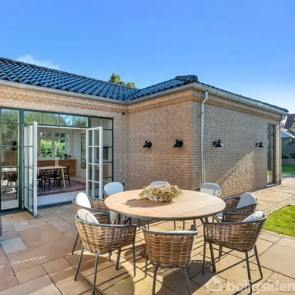 Rundt havebord med seks kurvestole står på terrasse foran murstenshus med åbne glasskydedøre. Indenfor ses et spisebord. Haven er grøn og solrig.