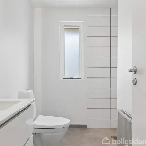Toilet i et moderne badeværelse; står langs væggen ved siden af en håndvask. Væggene er hvidmalede, og der er et smalt, frostet vindue bag toilettet.