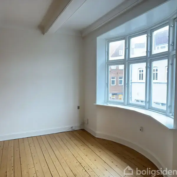 Tomt rum med trægulv og hvidmalede vægge, hviler under synlige loftsbjælker. Vindue mod gaden giver udsigt til bygninger udenfor. Ingen tekst er synlig.