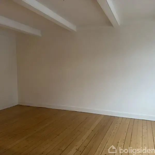 Et tomt rum med trægulv og hvidmalede vægge. Loftet har synlige bjælker, og der er god belysning. Minimalistisk og rent udtryk.