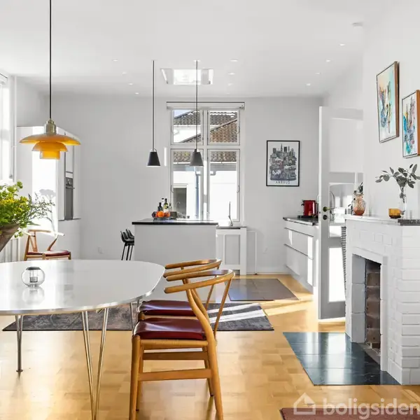 En lys, moderne stue med spisebord, omgivet af stilfulde træstole. Køkkenet er i baggrunden med hvide skabe, ovenlys, og indretningsdetaljer omfatter kunstværker og grønne planter.