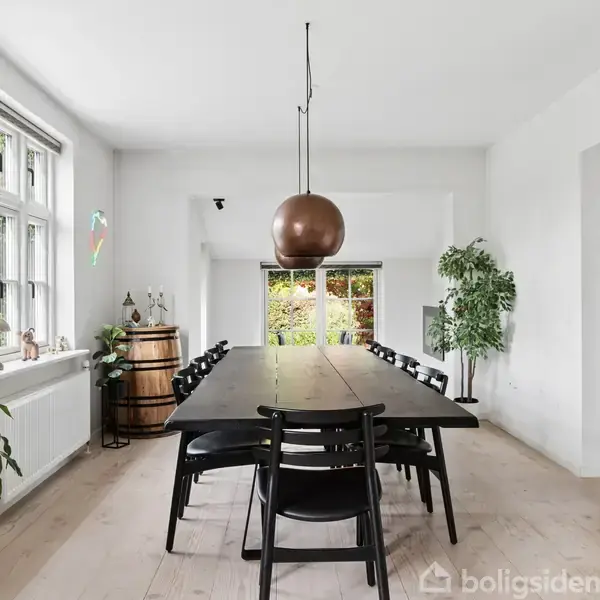 Et langt spisebord med sorte stole står i en lys spisestue. En kobberlampe hænger over bordet, der er omgivet af planter. Baggrund viser en trappe og en stor dør.
