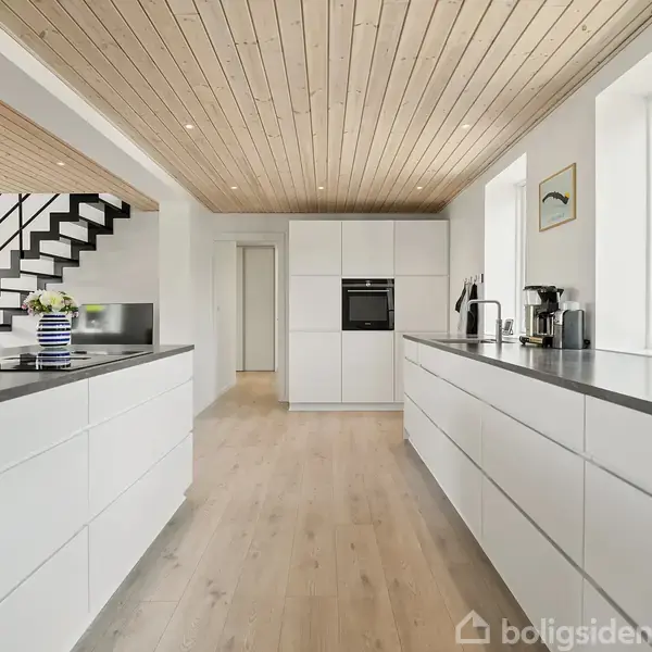 Hvidt køkken med moderne skabe. Skufferne åbne og kaffemaskine står på bordet. Store vinduer lader lys ind, og trappen til venstre fører til en overetage.