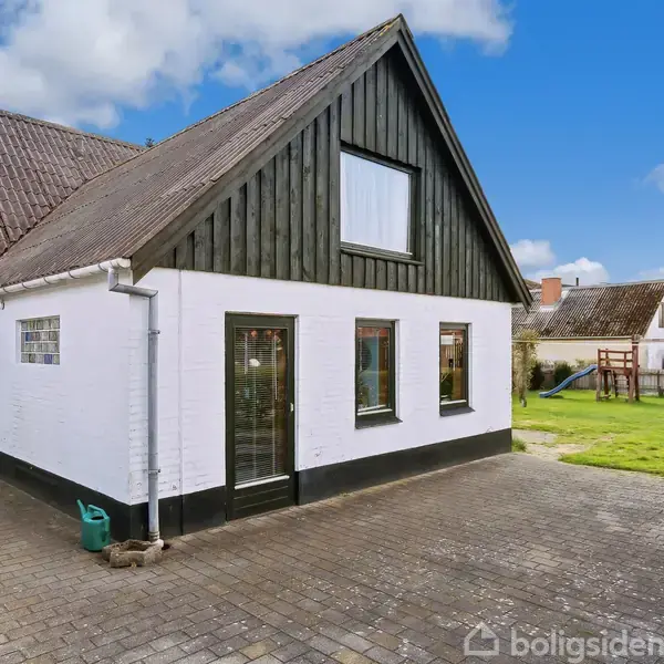 Et hvidt hus med mørkt tag står i en gårdhave. Der er flere vinduer og en glasdør. Husets baggrund er en græsplæne med et legehus og klare blå himmel.