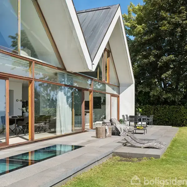 Et moderne hus har store glasvinduer og skråtag. Ved terrassen står solsenge og et bord med stole. Omgivet af en grøn have med udsigt til havet.