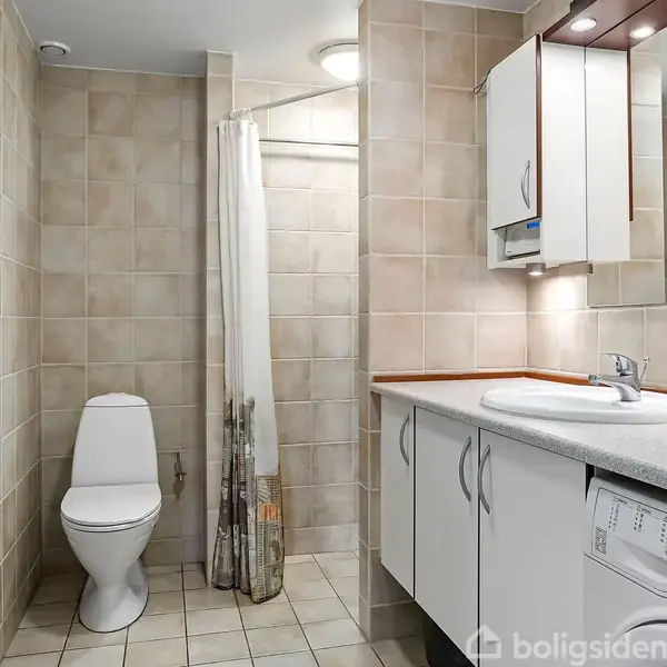 Toilet med lukket låg placeret ved siden af en bruser med forhæng; baderum omkranset af flisevægge og -gulv. Til højre ses en vask, spejlskab og vaskemaskine.