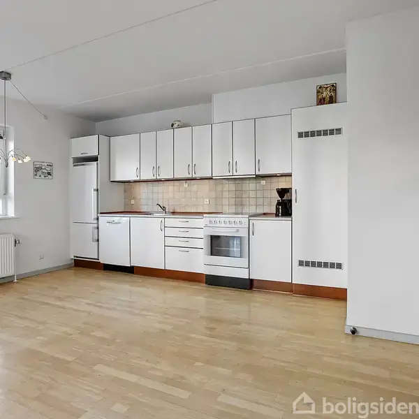 Et hvidt køkken i et åbent rum med trægulv. Skabe og apparater fylder bagvæggen. Et vindue og radiator ses til venstre, og en glasdør er til højre.