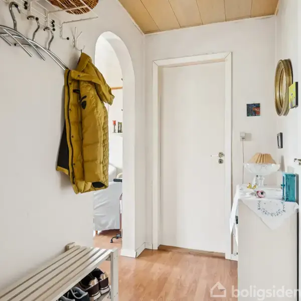 En entré har en gul jakke hængende fra en knagerække. En skohylde står under, mens en hvid kommode med pynt er til højre. Døren foran fører til et andet rum.