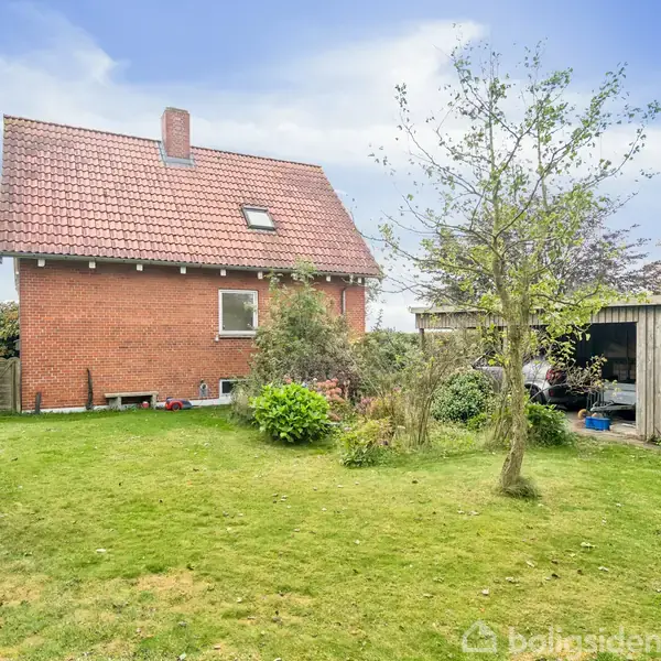 Et murstenshus med rødt tag står i en have med græs og et enkelt træ. Til højre er en trægarage. Himlen er blå med spredte skyer.