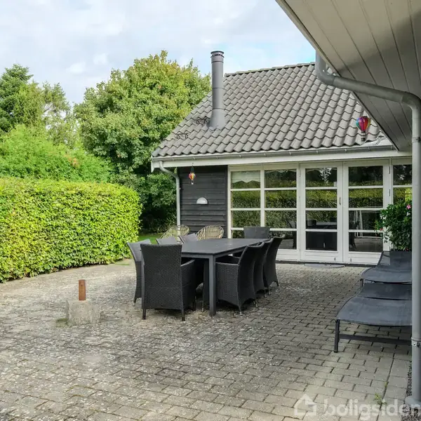 Terrasse med havemøbler står på flisebelagt areal foran et moderne hus med skiferet tag. Omkranset af grønne hække og træer, skaber en privat og hyggelig atmosfære.