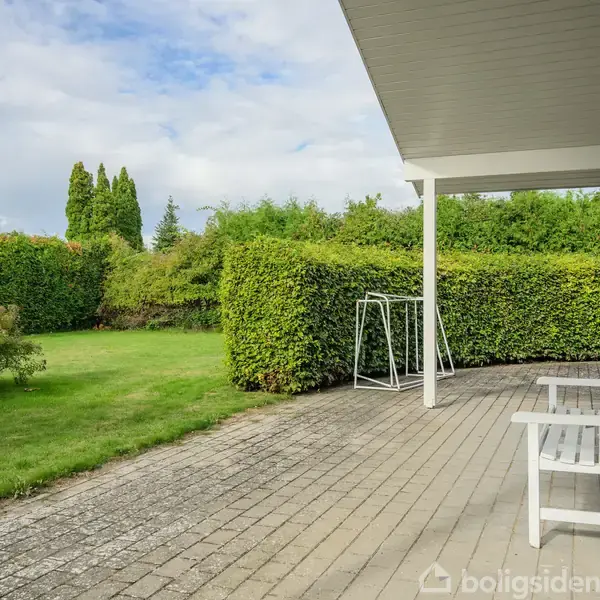 En træbænk står stille på en terrasse med fliser ved et hus. I baggrunden er velplejede hække og en grøn græsplæne under en delvis skyet himmel.