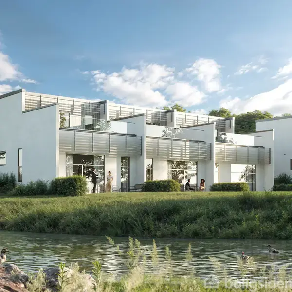 Rækkehuse står stille ved en sø med frodig vegetation i forgrunden. Sollys skinner på bygningens hvide facader, omgivet af grønne træer under en blå skyfri himmel.