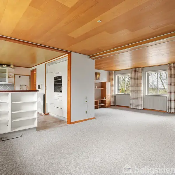 Stue med åbent køkken viser et lyst rum med trægulv og lofter. Køkkenet til venstre har hvide hylder. Store vinduer til højre giver udsigt til en have.