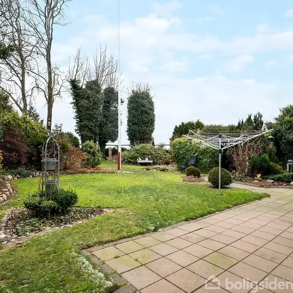 Græsplæne med metallisk tørrestativ omgivet af en flisebelagt terrasse. Haven har buske og træer i baggrunden under en lys, skyet himmel.
