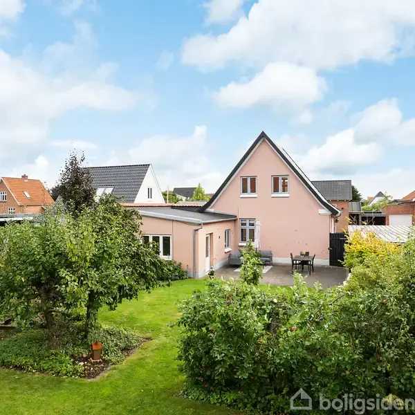 Et lyserødt hus med spidst tag står stille i en velholdt have med grønne buske og træer, omgivet af andre huse under en blå himmel med skyer.