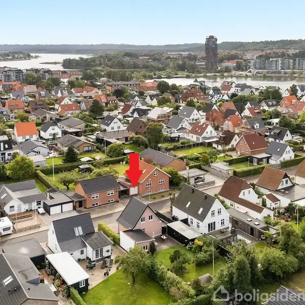 Rødt murstenshus peget ud af en rød pil i et boligområde med mange huse omkring. I baggrunden ses en flod og flere bygninger.