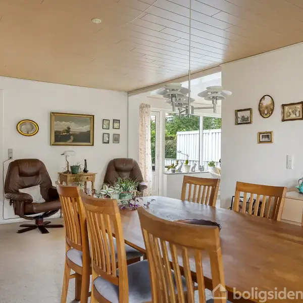 En spisestue med træstole står omkring et ovalt bord. To lænestole er placeret ved væggen, udsmykket med malerier. Vinduet giver udsigt til en have.