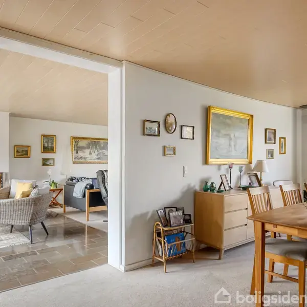 Spisebord med stole står i en spisestue ved siden af en kommode med billedrammer. I baggrunden ses en stue med sofaer, lænestole og pejs.