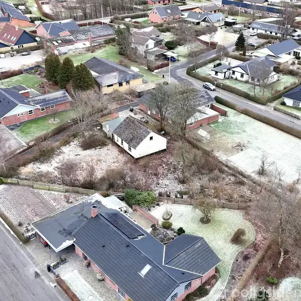 En hvid bygning med stråtækt tag står stille omgivet af træer og græs i et parcelhuskvarter med mange andre huse. Asfalterede veje løber gennem området.