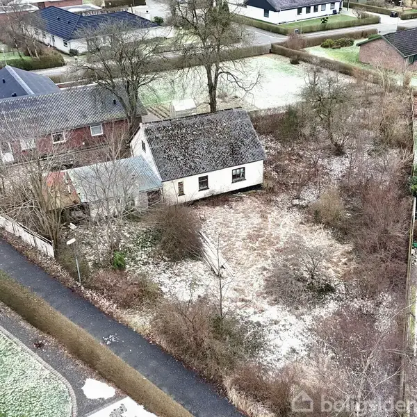 Et ældre, forfaldent hus står stille i en tilgroet have omkranset af buske og træer. Omkring huset ligger en stille villavej med flere andre parcelhuse.