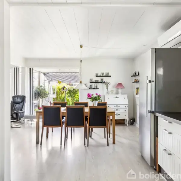 Spisebord med seks stole står i et lyst, moderne køkken/alrum med hvide skabe og glasløse vægge, som giver udsigt til en have. En køleskab og en kommode er også synlige.