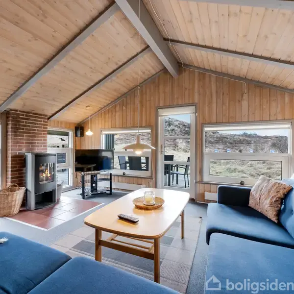 Blå sofaer står i et stueområde med trævægge og skråt loft. En brændeovn er midtpunktet, omgivet af store vinduer der viser en landskabelig udsigt. Afslappet atmosfære med moderne indretning.
