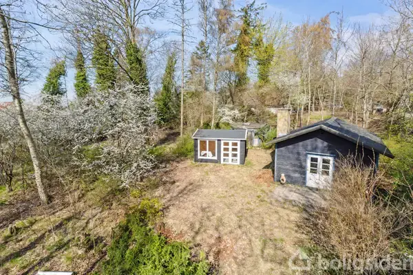 To små huse står i en skovklædt have omgivet af træer. Et hus har en mørk træfacade, mens det andet er lysere med glasdøre.