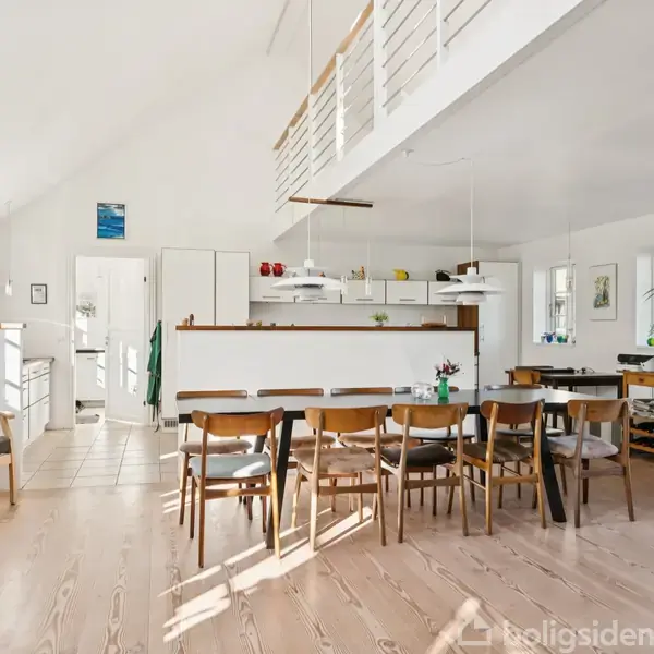 Lys spisestue med seks stole omkring et bord. Køkkenet i baggrunden har hvide skabe. Rummet har lyst trægulv, loft til kip og et moderne interiør.