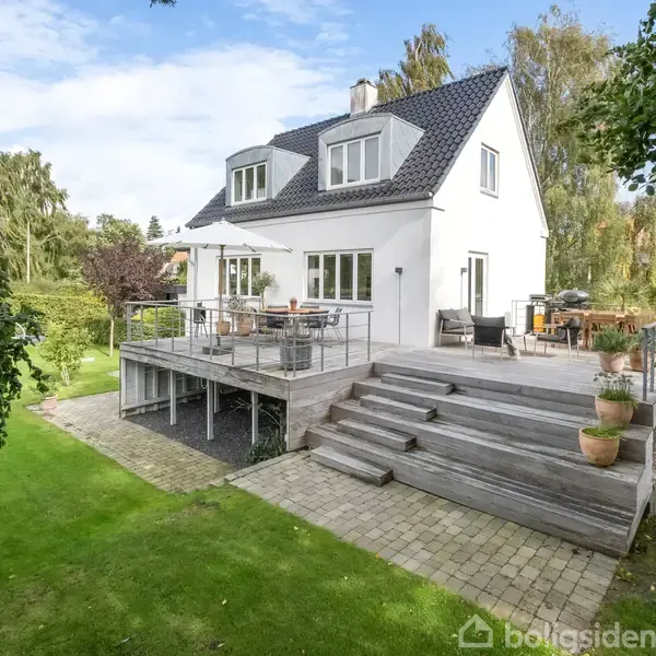 Hus med hvid facade står i fokus, udstyret med stor træterrasse fyldt med havemøbler. Græsplænen omgiver ejendommen, omkranset af høje træer og buske under en blå himmel.