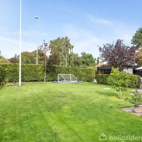 En græsplæne med et fodboldmål står foran en hæk. Til højre ses en terrasse med havemøbler ved siden af et hus. Himlen er blå med enkelte skyer.