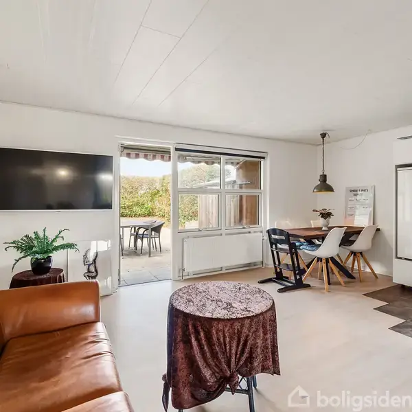En stue har en brun lædersofa og et rundt bord med dug. Et fladskærms-TV hænger på væggen. Til højre er en spisestue med bord og stole samt et åbent køkken. En dør fører ud til en terrasse med havemøbler.