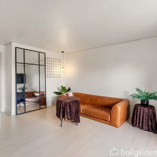 En brun lædersofa står mod en hvid væg med en væg-tv til højre. Små borde med planter og spejle til venstre skaber en hyggelig atmosfære i stuen.