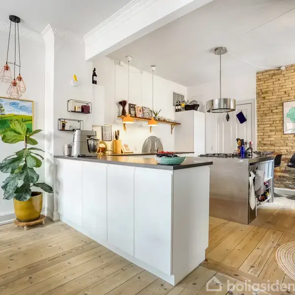 Køkken med hvidt køkkenbord og hængende lamper, omgivet af planter og kunst på væggen. Trægulve og murstensvæg bagved. Ingen tekst til stede.