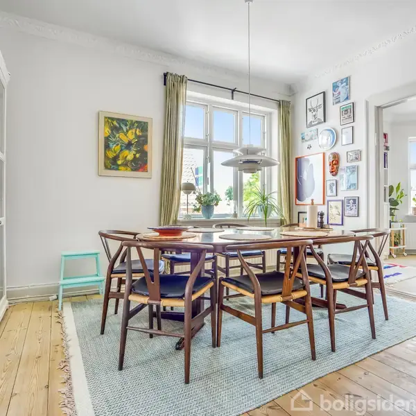 Et spisebord med stole omkring det står på et lyst tæppe i en stue. Rummet har trægulve, hvide vægge, kunstværker, og et vindue med grønne planter på vindueskarmen.