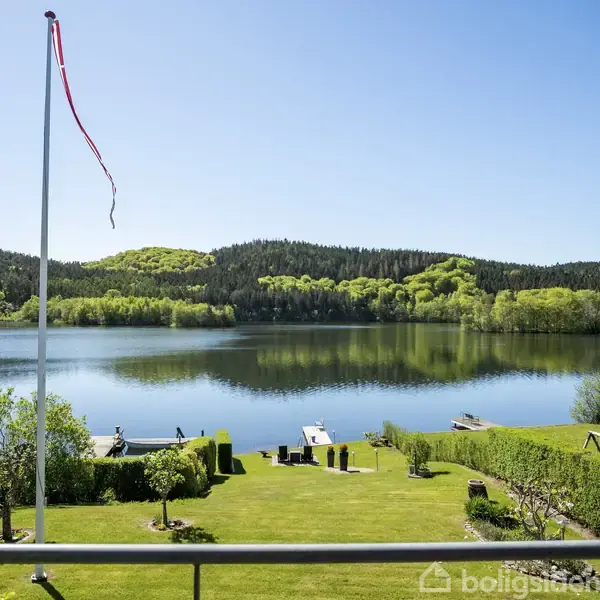 En glasdør åbner ud mod en balkon, hvor en flettet stol står. Udsigten viser en sø med skovklædte bakker i baggrunden under en klar blå himmel.
