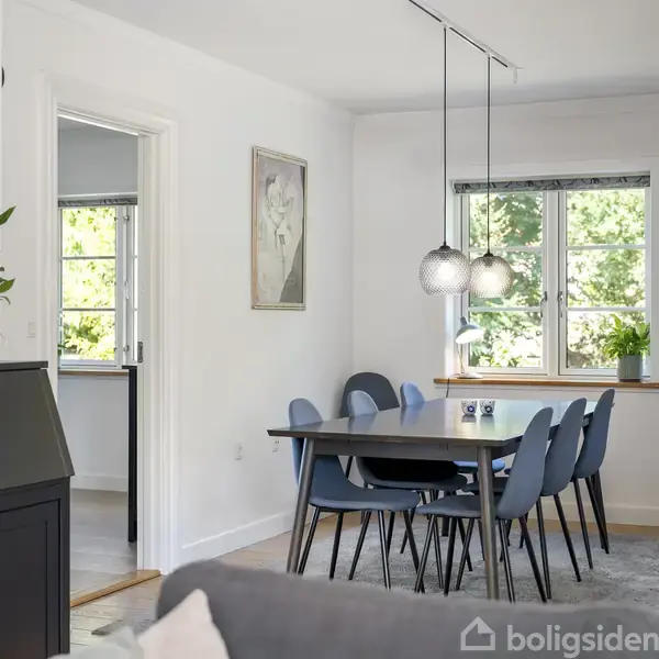 Spisebord med blå stole står ved vindue. Over bordet hænger tre lamper. Grønne træer kan ses udenfor. På væggen hænger et maleri. En plante står på et skab.