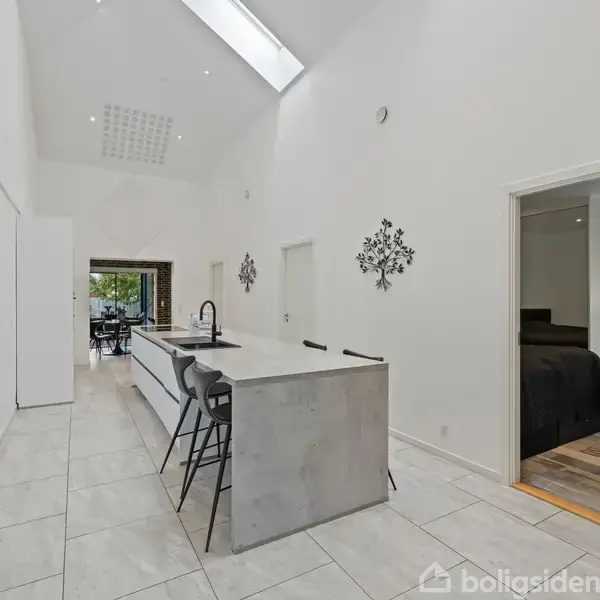 Køkkenø med barstole står i en lys, moderne gang med højt til loftet. Til højre er en åben dør, der leder ind til et soveværelse.