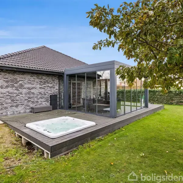 Hus med muret ydervæg, har glasudestue ved siden af en terrasse med indbygget jacuzzi. Omgivet af græsplæne og et træ. Himlen er blå.
