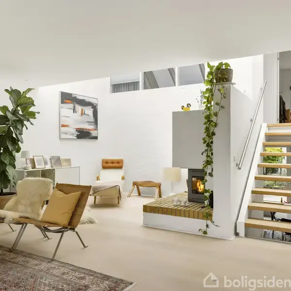 En moderne stue med lyse vægge og minimalistisk indretning. En åben pejs er centreret i rummet; en trappe fører til et højere niveau. Grønne planter dekorerer rummet.
