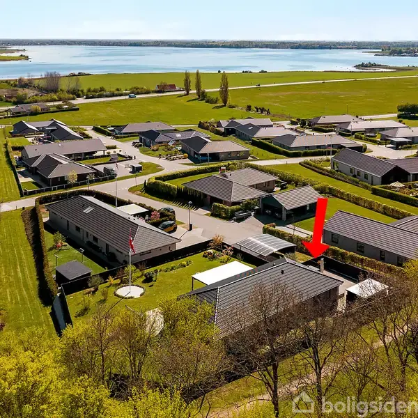 Hus markeres med rød pil, set fra oven i et kvarter med mange ens huse. Omgivet af grønne plæner og træer, med udsigt til en sø i baggrunden.