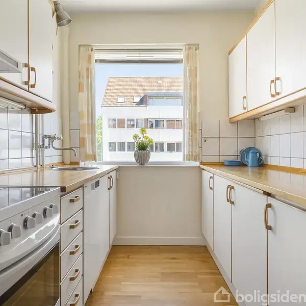 Et lyst køkken med træskabe og hvide fliser. En moderne ovn står til venstre. I midten er et vindue med en grøn plante i en vase. Udenfor ses et rødt tag.
