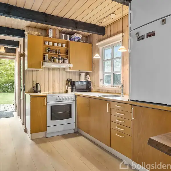 Køkken med træpaneler og sorte loftsbjælker, indeholder et køleskab, komfur, og mikrobølgeovn, omgivet af lyse træskabe. Et vindue giver udsigt til en grøn have.