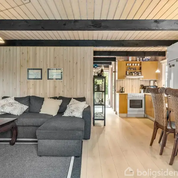 En grå sofa med hjørnemodul står i et stueområde med trægulv og lyse træklinker. Ved siden af er et spisebord med kurvestole, og i baggrunden ses et åbent køkken.