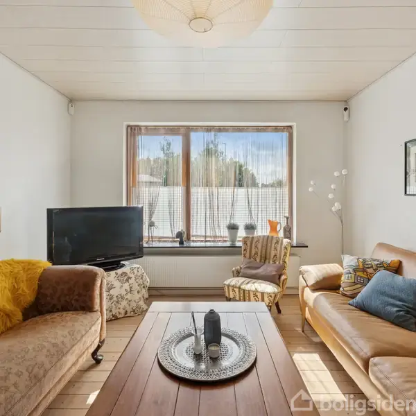 En stue med to sofaer og et sofabord, en lampe, og en fladskærms-tv. Store vinduer lader sollys ind, mens kunst og dekorative genstande pryder væggene og vindueskarmen.