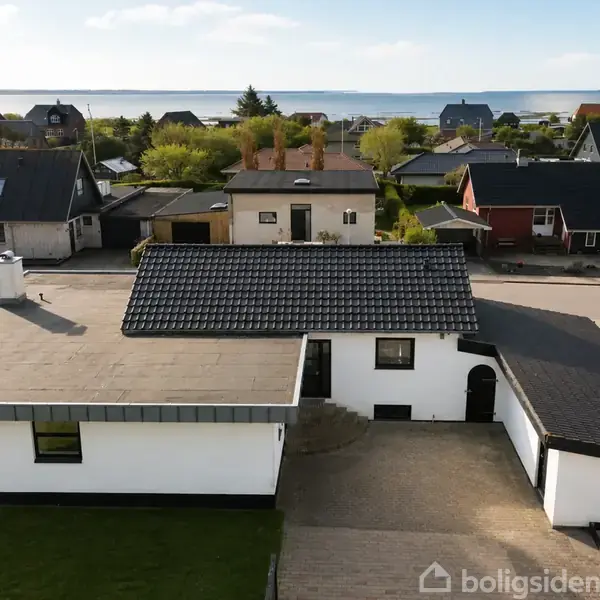 En hvid, moderne villa med fladt tag og indkørsel står forrest. I baggrunden ligger flere huse og en havudsigt.