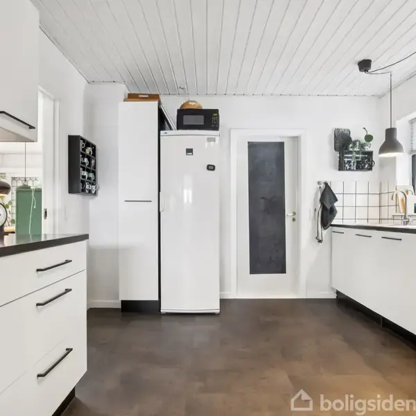 Et moderne køkken med hvide skabe og sorte bordplader. Et køleskab står ved siden af en dør. Vinduer giver naturligt lys, og en vægt samt lamper tilføjer detaljer.