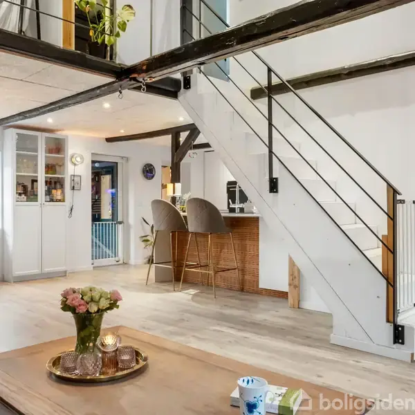 Træbord med blomster og lys står i en moderne stue, hvor en hvid trappe med sorte gelændere leder op til en åben førstesal. Køkken ses i baggrunden med barstole.